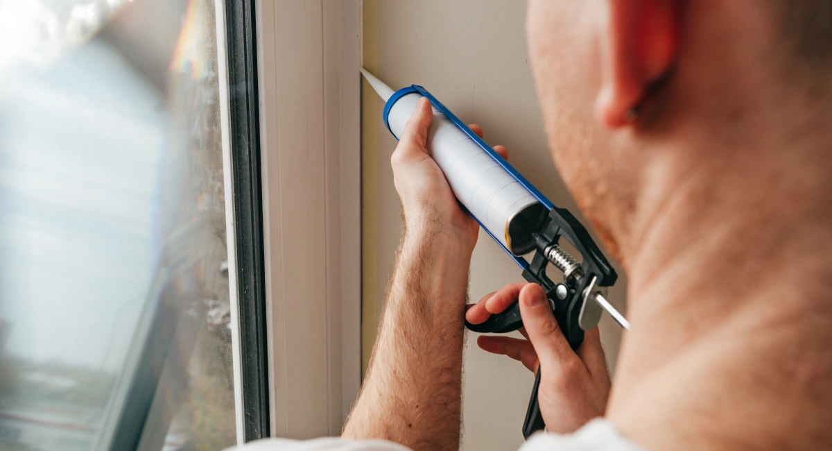man wearing overalls sealing cracks between window and trim