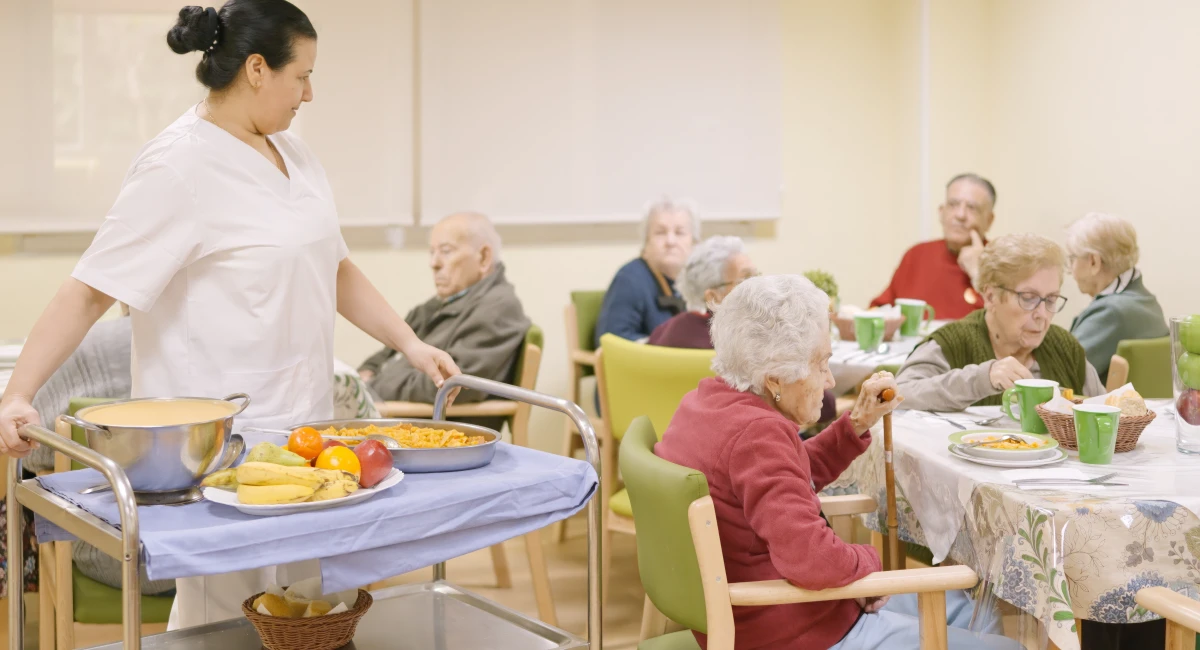 Dining room of care home filled with residents and worker handing out dinner